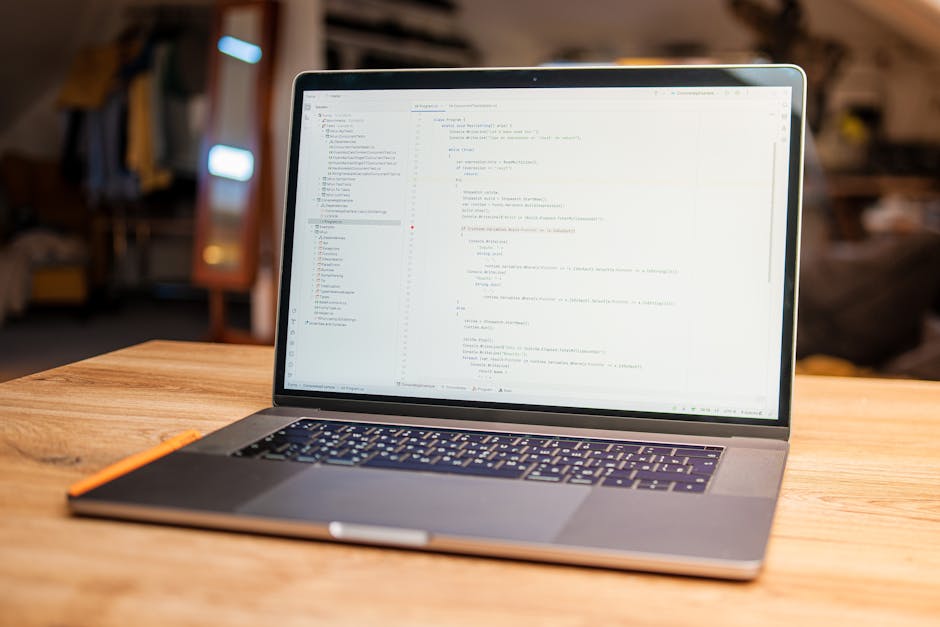 A close-up view of a modern white desk in a well-lit room, featuring a silver laptop displaying lines of colourful code on its screen, a white smartphone placed on the laptop's trackpad, an open blank